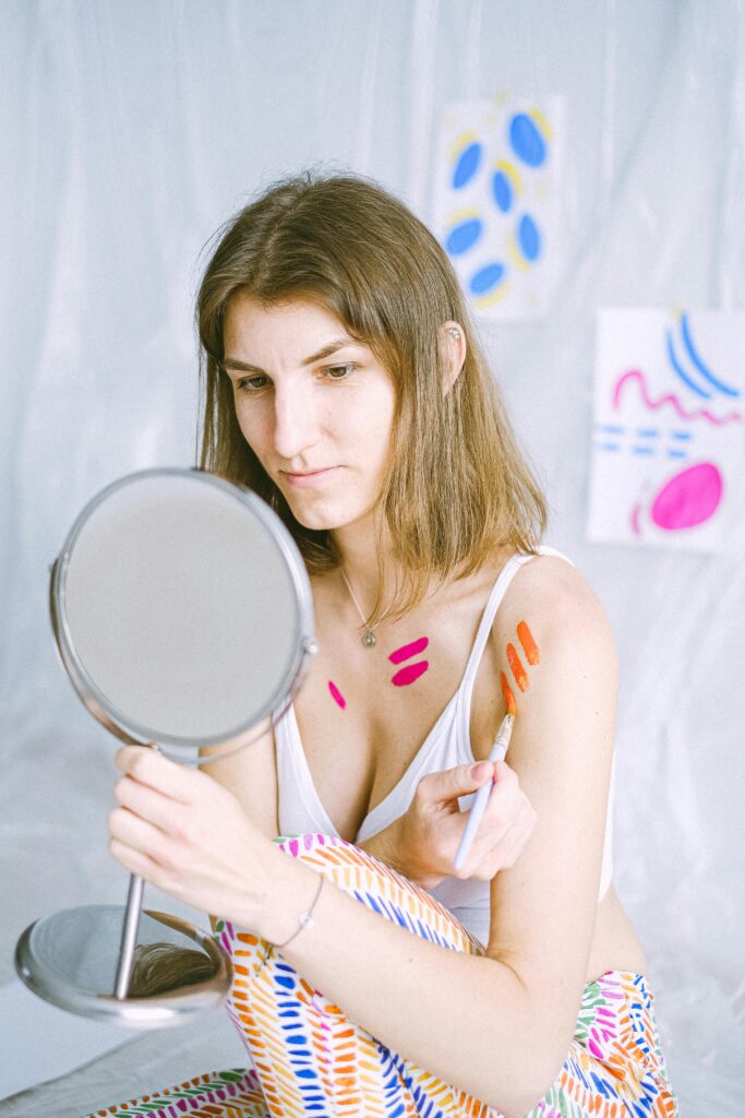 Woman applying body paint while looking in mirror during creative workshop. Selbstfürsorge für Frauen*