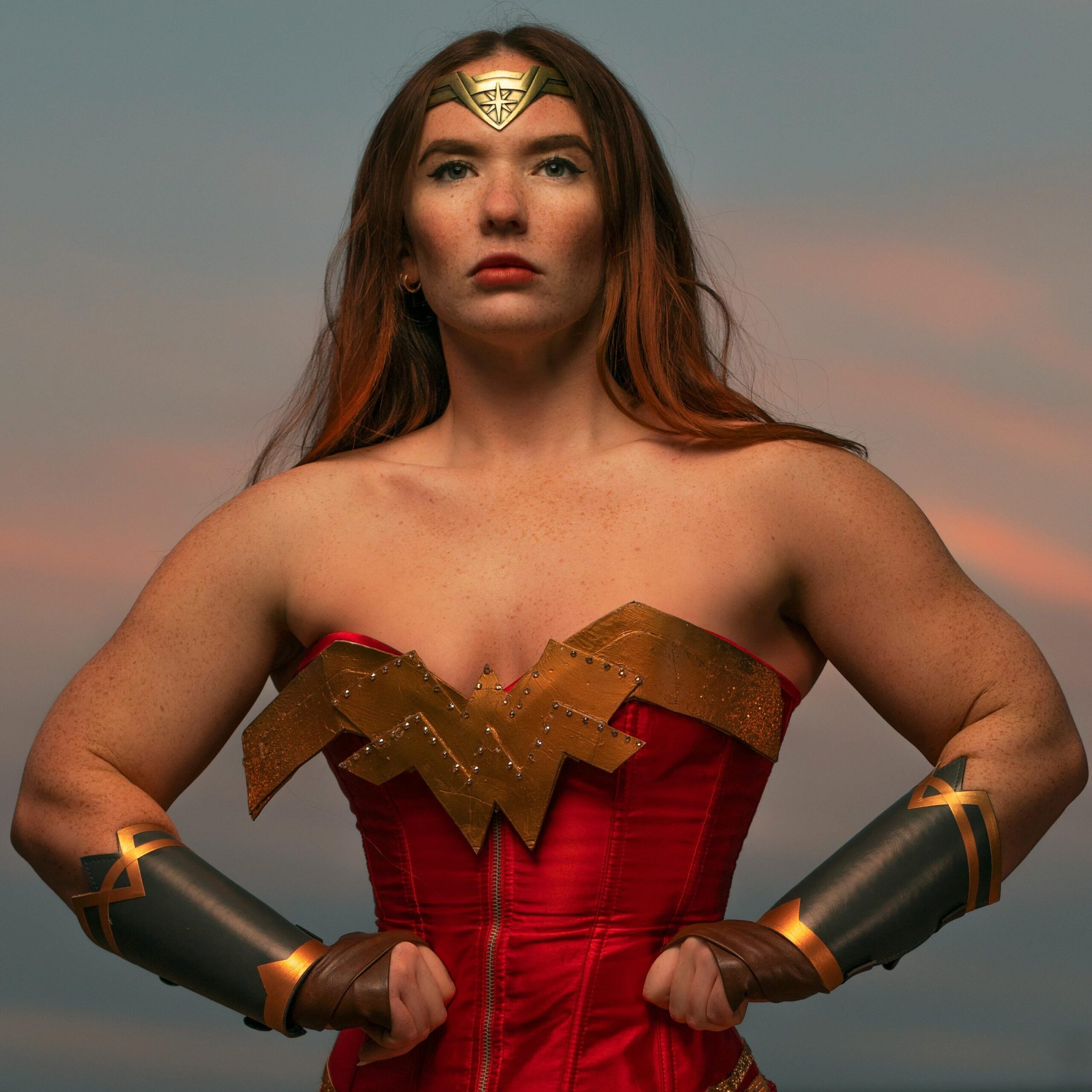 A strong woman dressed as a superhero posing with confidence outdoors during sunset.